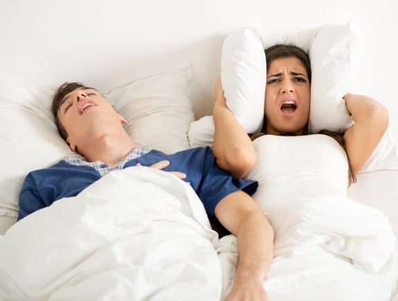 Woman in bed angrily covering her ears with pillow while man with sleep apnea snores next to her
